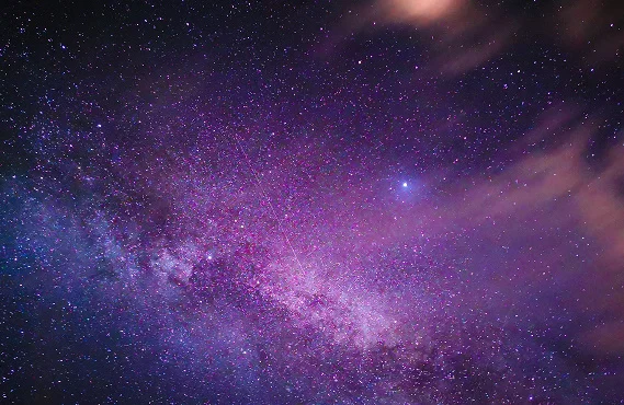 Person sitting on a mountain cliff beneath a colorful galaxy filled with stars, glowing nebula clouds, and a breathtaking cosmic night sky.